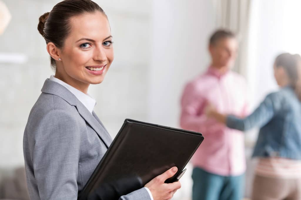 Portrait Of Smiling Young Female Realtor Holding Document With Clients In Background